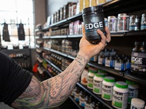 close up of a males arm and hand holding pre workout supplement with a rack of supplements in the background